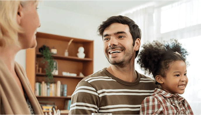 Father holding daughter in lap and smiling at woman