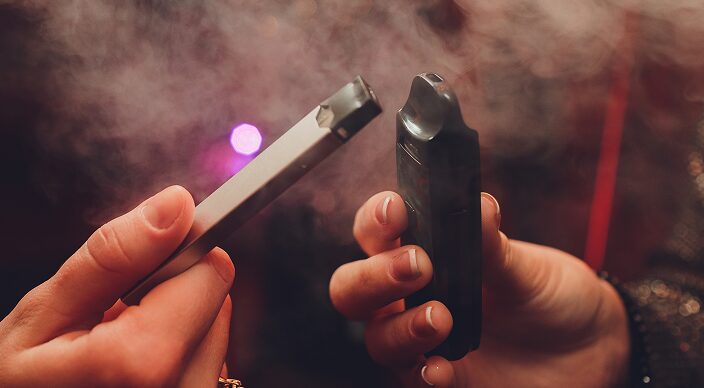 Two people holding two different kinds of vapes surrounded by smoke
